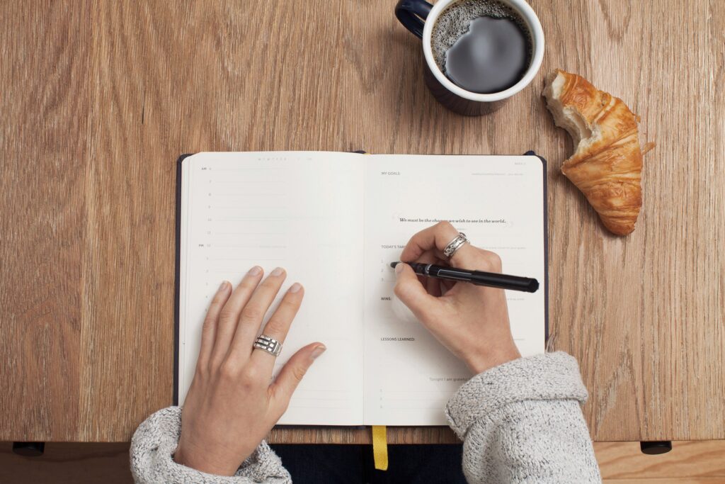 Aus der Vogelperspektive ist ein Schreibtisch fotografiert auf dem rechts oben eine Tasse mit Kaffee steht und daneben ein angebissenes Croissant liegt. Im Zentrum des Tisches befinden sich ein Bullet Journal sowie zwei Unterarme einer Frau. Die Frau schreibt in das Tagebuch.