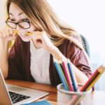 Frau mit Brille und langen dunklen Haaren sitzt vor ihrem Laptop und sieht erwartungsvoll auf den Bildschirm.