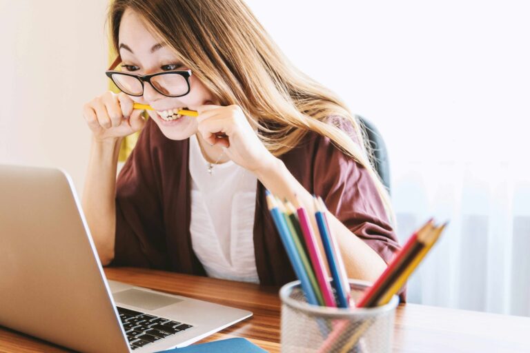 Frau mit Brille und langen dunklen Haaren sitzt vor ihrem Laptop und sieht erwartungsvoll auf den Bildschirm.